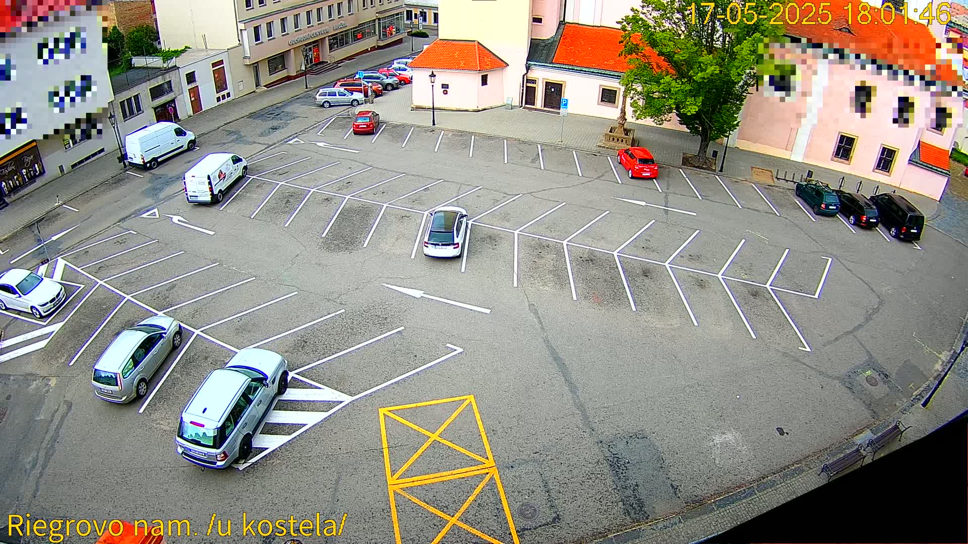 An empty parking lot with several cars parked in it, surrounded by buildings and a tree, on a sunny day.