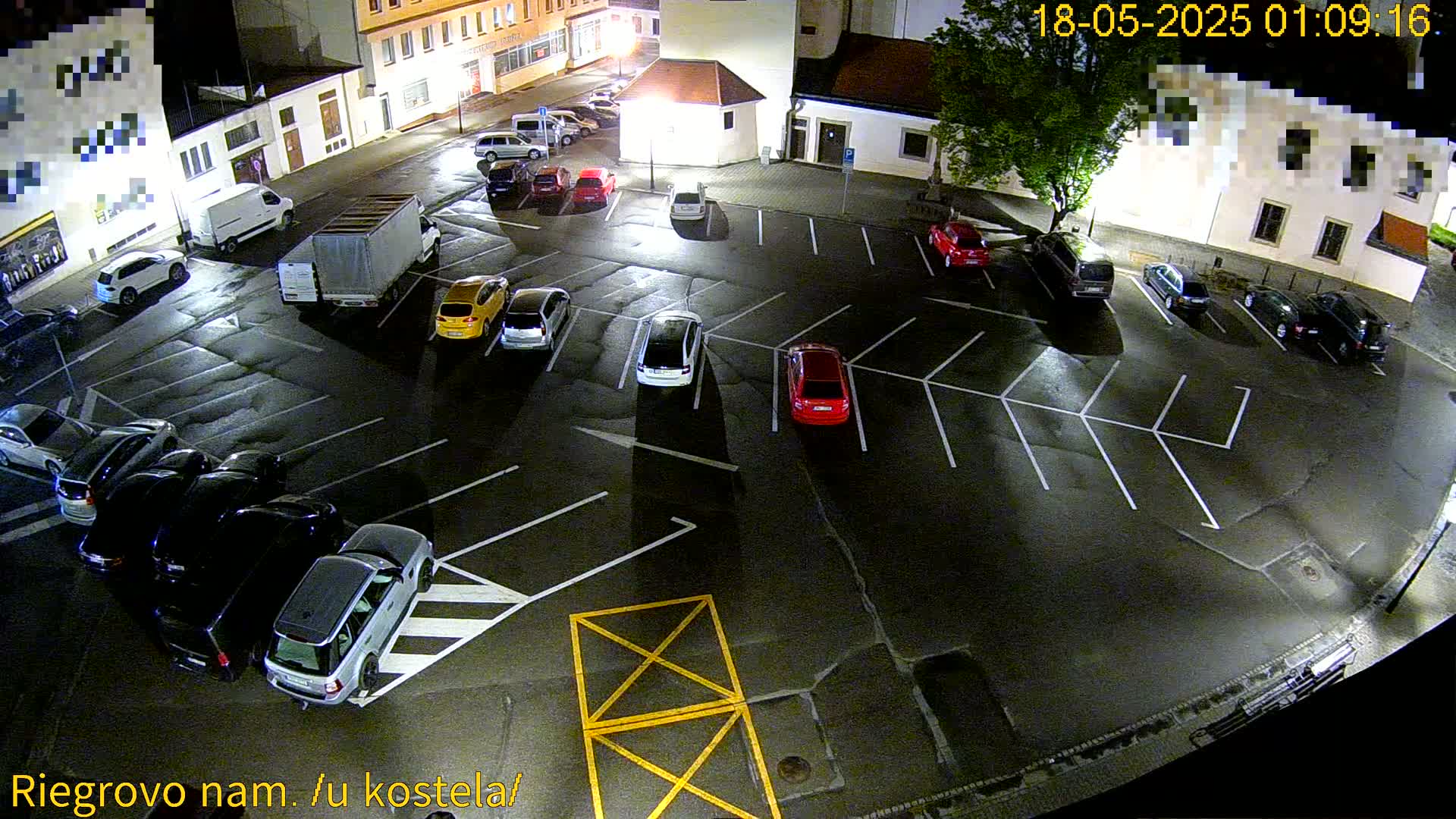 A nighttime, overhead view of a mostly empty parking lot with several cars and vans parked, illuminated by streetlights.