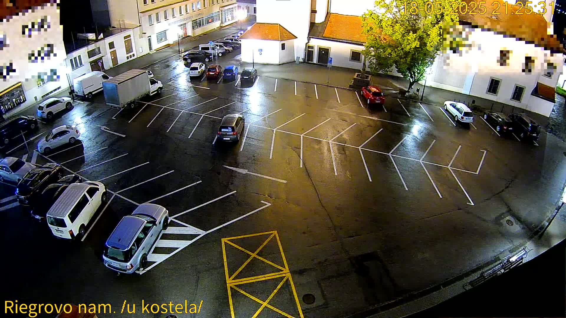 A wet, nighttime parking lot is sparsely populated with several cars and vans parked in designated spaces near a church.