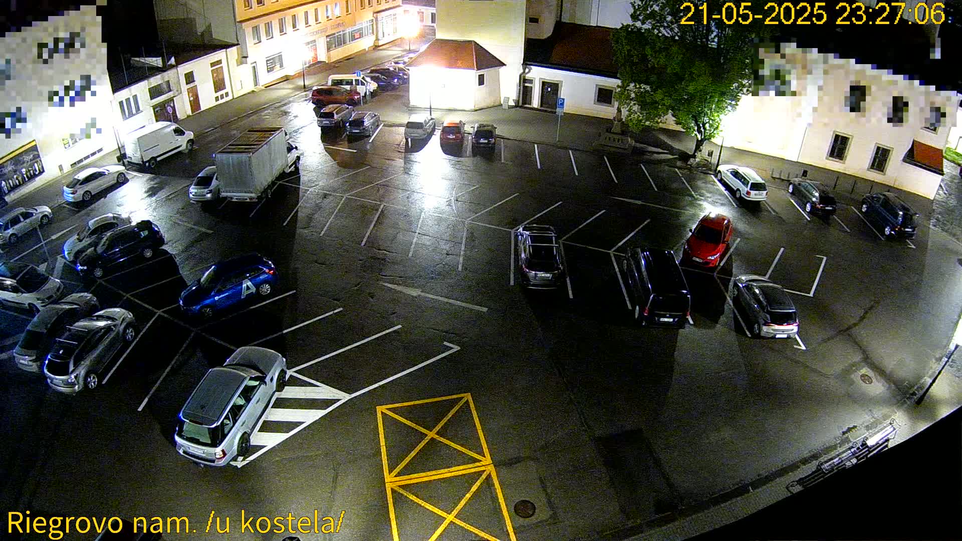 A wet, nighttime parking lot is mostly filled with cars, with a few empty spaces, and a large truck parked near the edge.