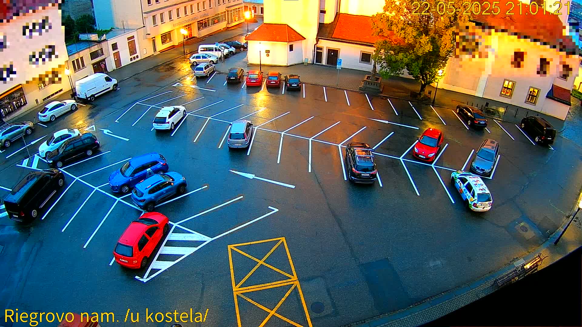 A wet, asphalt parking lot at dusk is filled with various cars parked in designated spaces, with several buildings visible in the background.