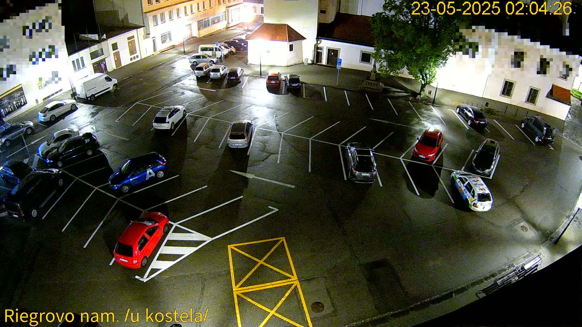 A wet, nighttime parking lot is filled with numerous cars parked in designated spaces, under dim lighting.