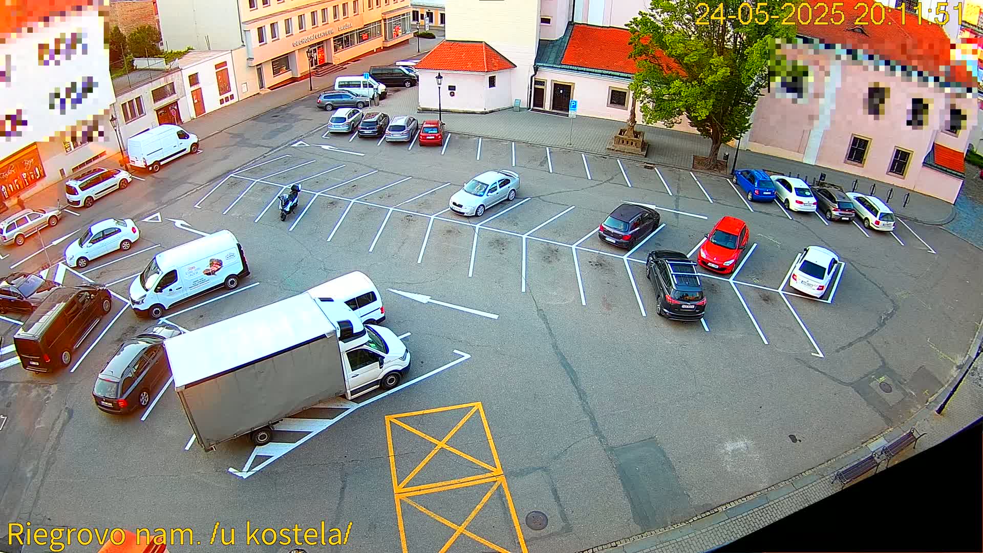 An aerial view shows a parking lot with numerous cars and vans parked, a large truck maneuvering, and a person on a scooter; the weather appears to be clear and daytime.