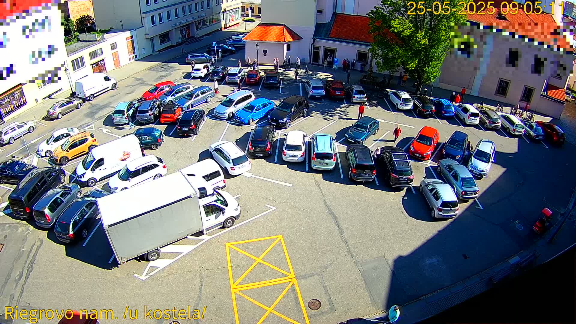A sunny outdoor parking lot is filled with numerous cars, a large white box truck, and a few pedestrians.