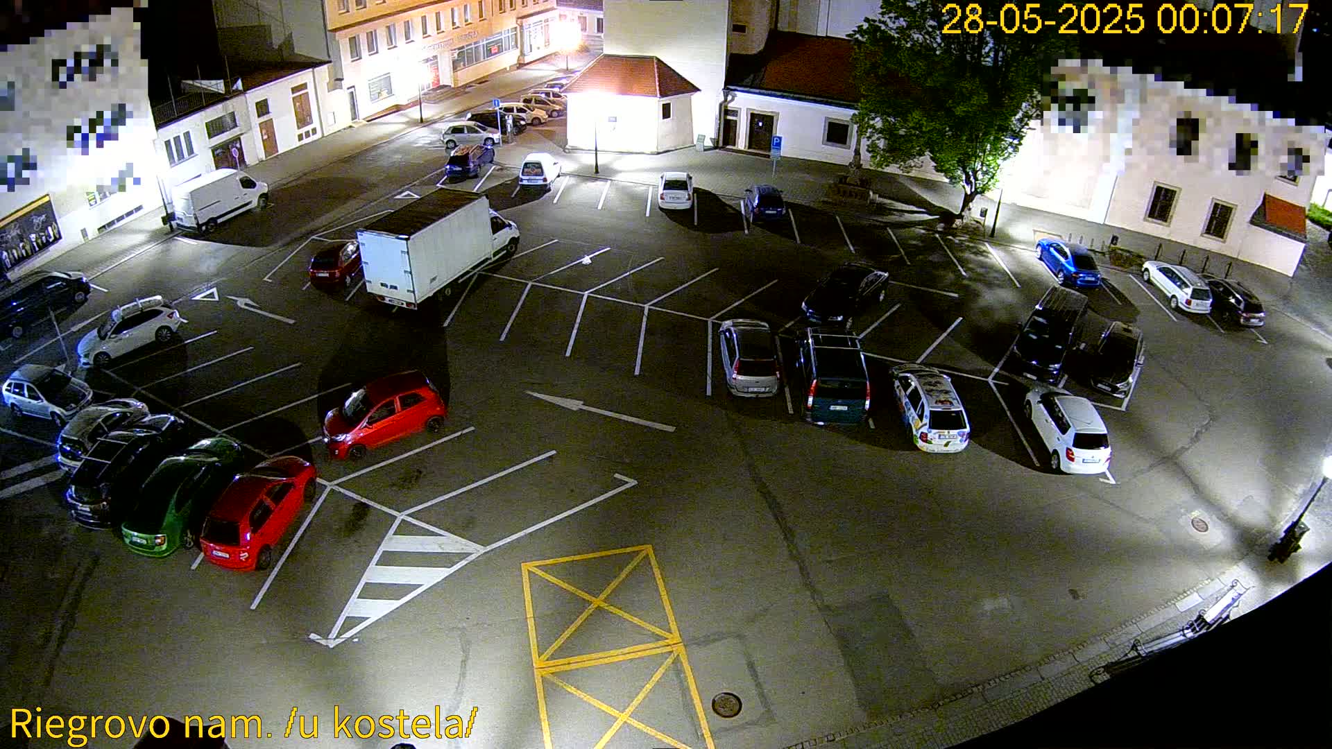 A nighttime overhead view of a mostly empty parking lot with several cars parked, a white delivery truck, and surrounding buildings.