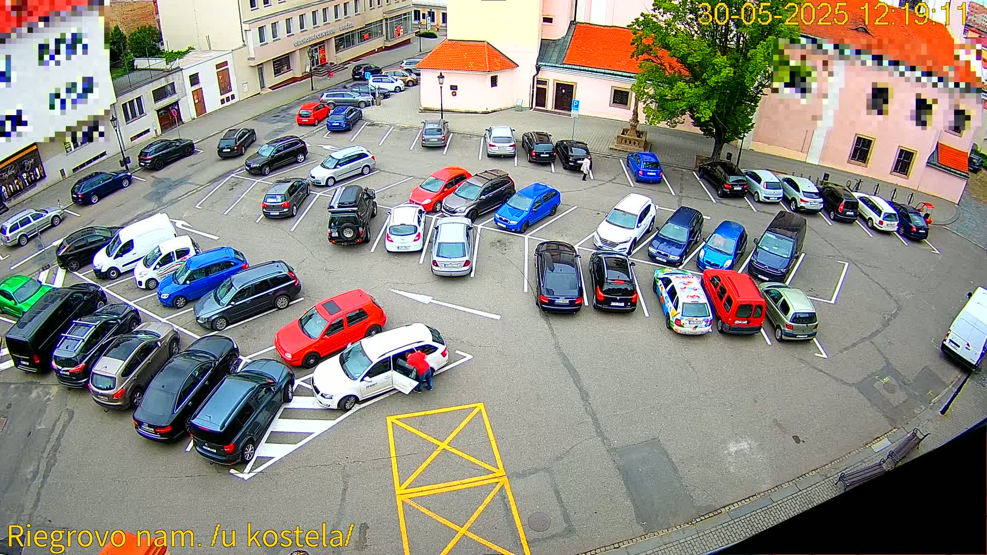 An aerial view shows a person loading items into a white station wagon parked in a mostly full parking lot, surrounded by buildings on a partly sunny day.