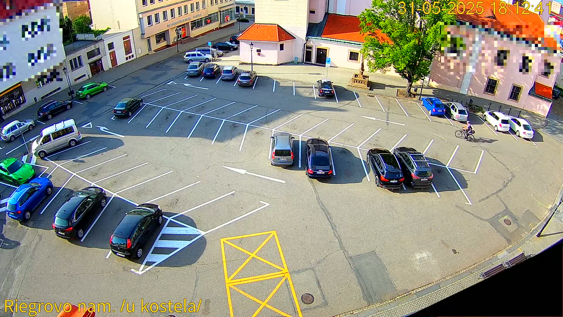 A mostly empty parking lot with several cars parked, a person riding a bicycle, and buildings in the background on a sunny day.
