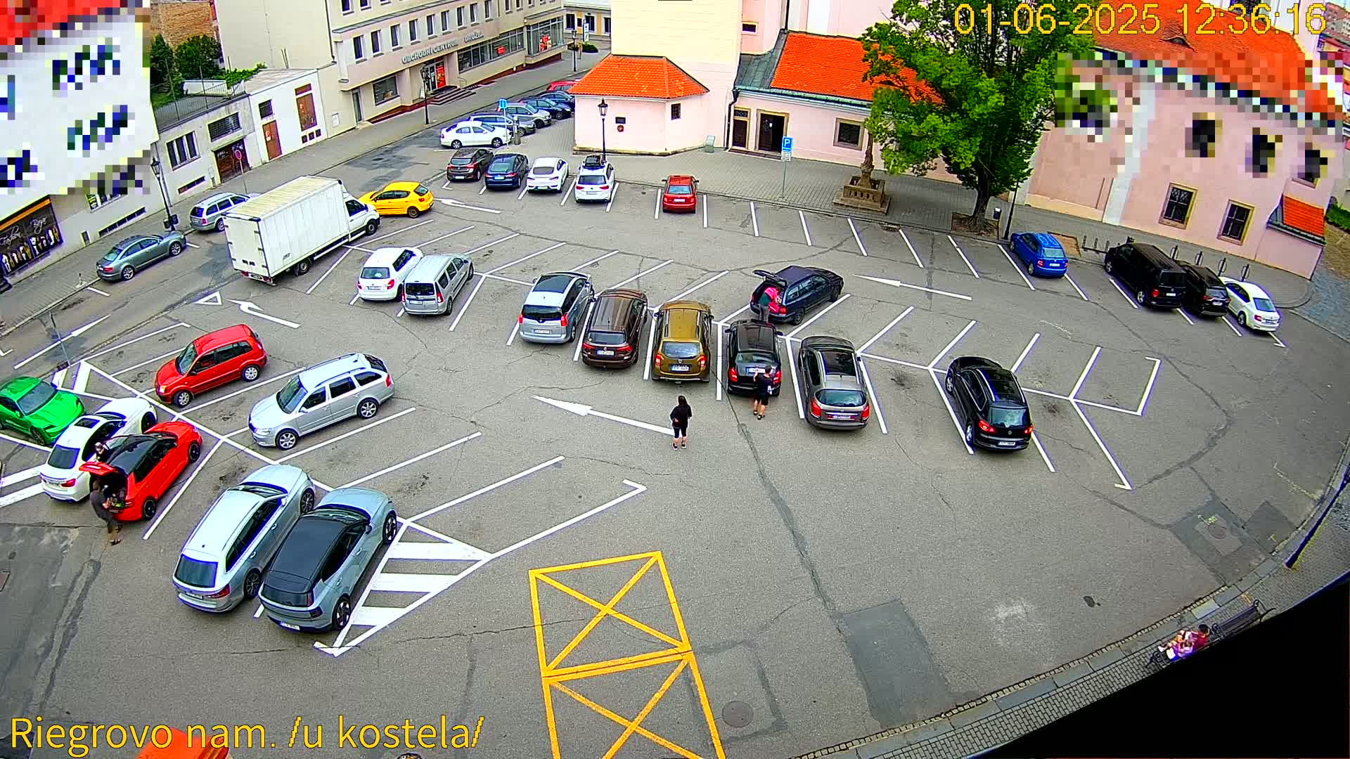 A mostly full parking lot with numerous cars parked in marked spaces, and a few people standing near some of the vehicles, under a partly sunny sky.