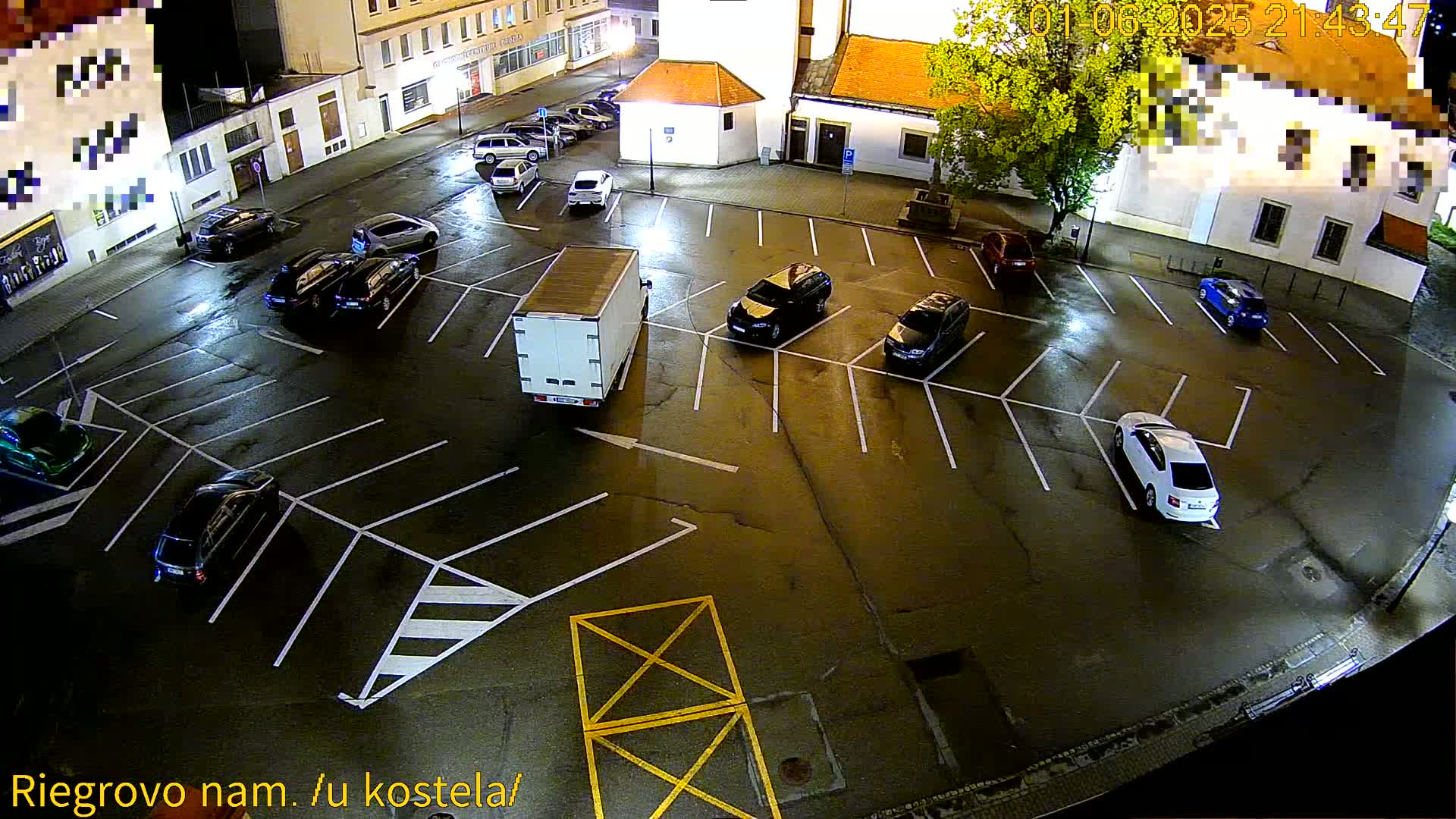 A nighttime overhead view of a mostly empty parking lot with several cars parked and a white box truck, under wet conditions.