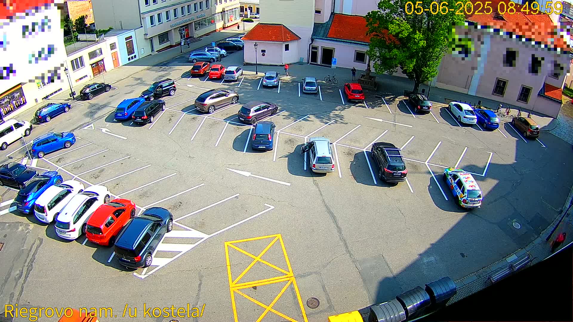 A sunny day shows a mostly full parking lot with various cars parked in spaces, surrounded by buildings.