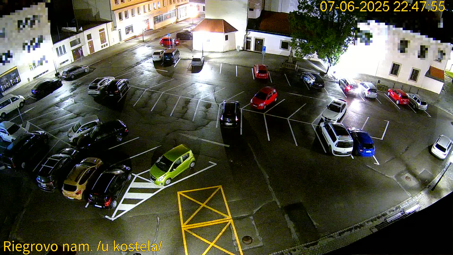A nighttime aerial view of a mostly empty parking lot with several cars parked in it under wet conditions.