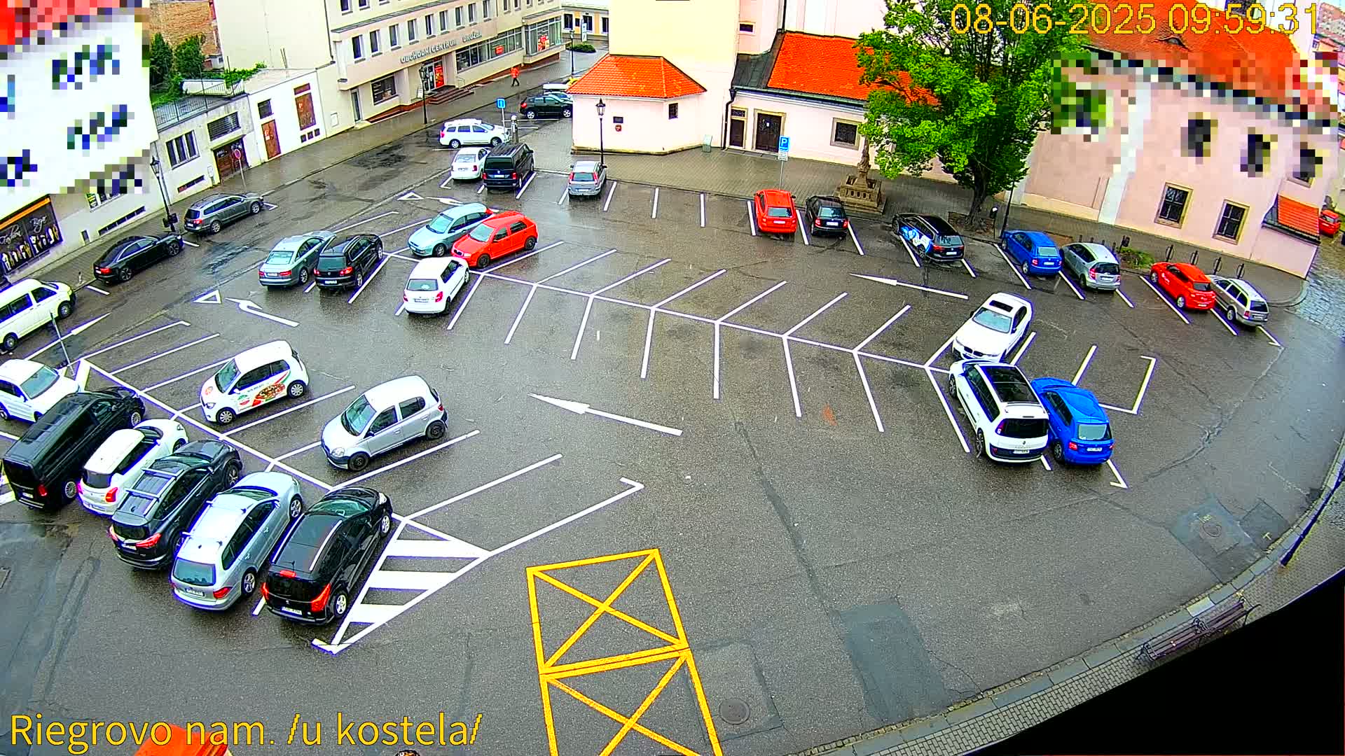 A wet parking lot filled with numerous cars is surrounded by buildings on a cloudy day.