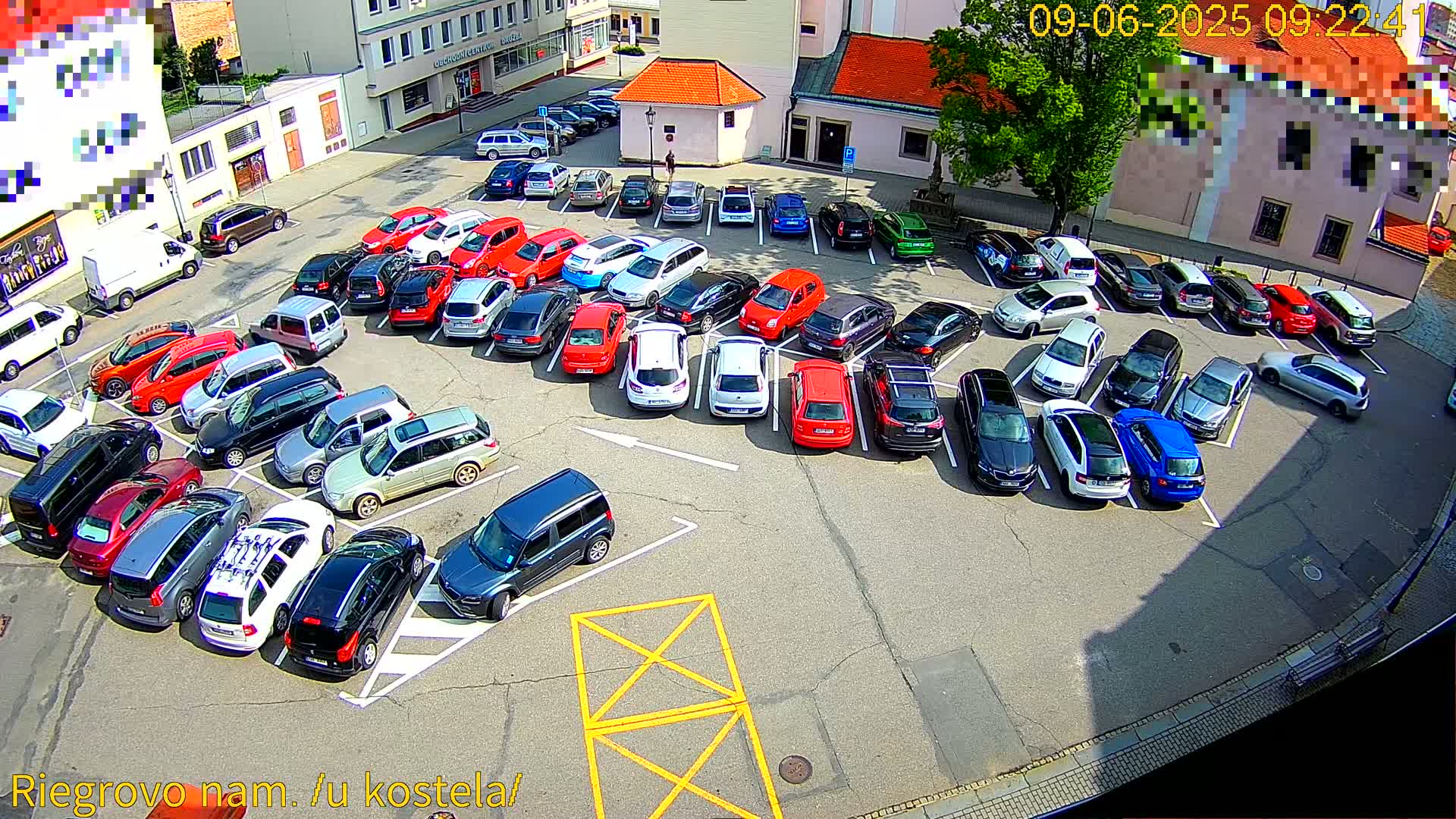 A sunny day reveals a full parking lot with numerous cars parked in organized rows, surrounding a small plaza near buildings.