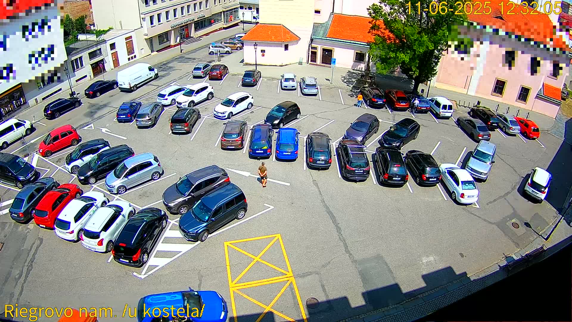 A man stands in a sunny parking lot filled with numerous cars, many parked in rows.