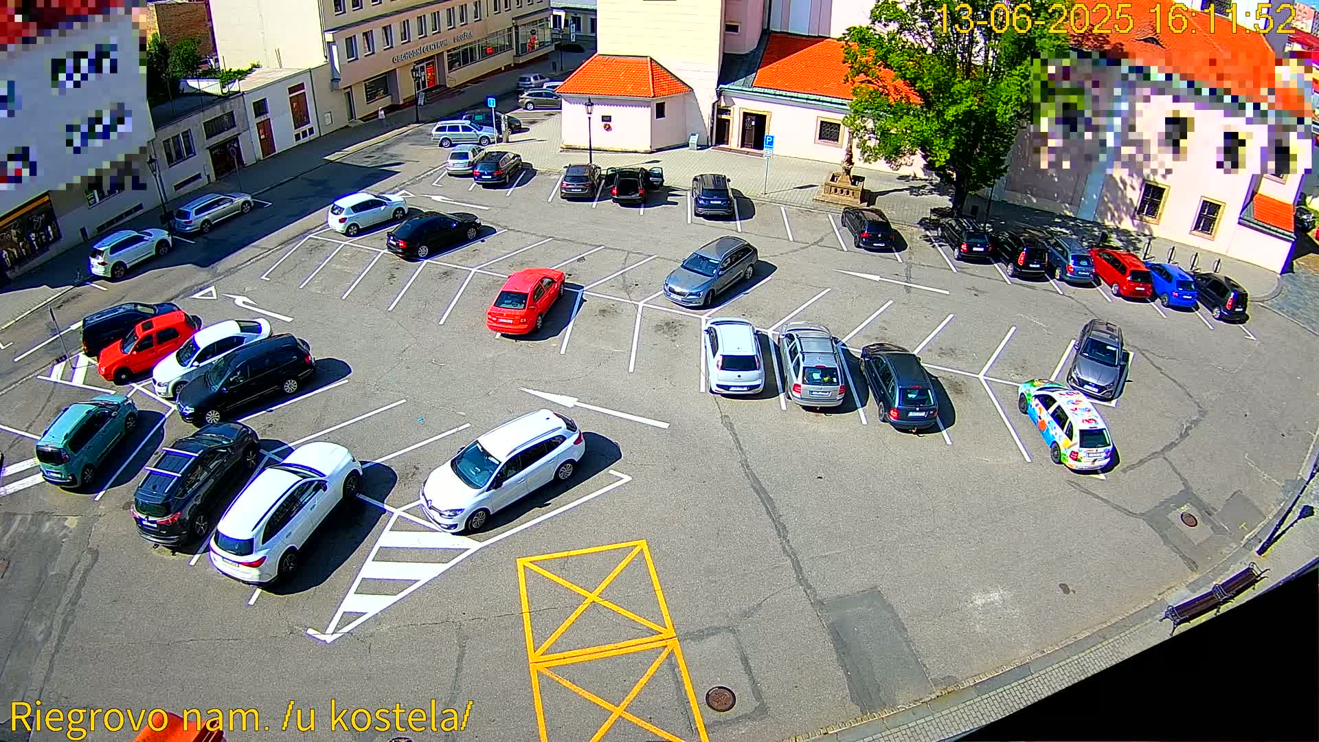 An aerial view shows a sunny parking lot filled with cars surrounding a church, with a few cars parked on a street adjacent to the lot.
