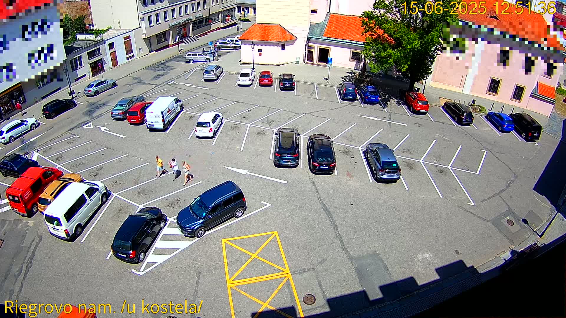 A sunny day shows a parking lot filled with cars, and three people walking near a row of parked vehicles.