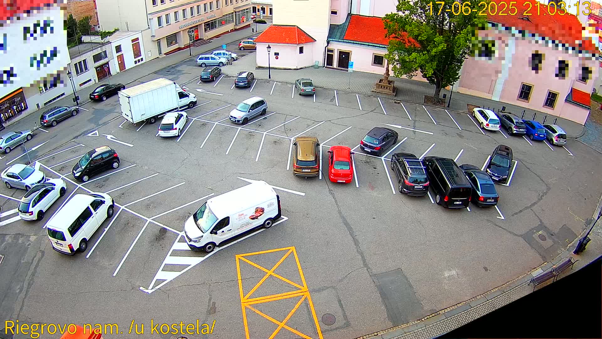 An aerial view of a parking lot with numerous cars and vans parked in a town square on a partly cloudy day.