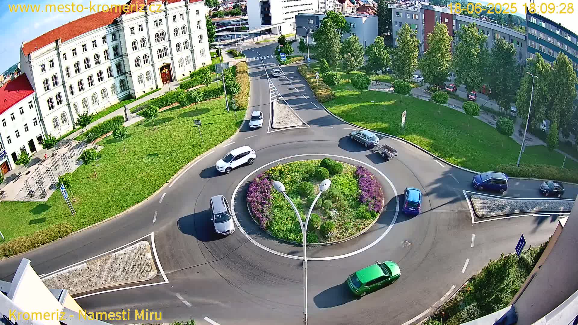 An aerial view of a roundabout surrounded by green lawns and buildings, with several cars navigating the circle on a sunny day.