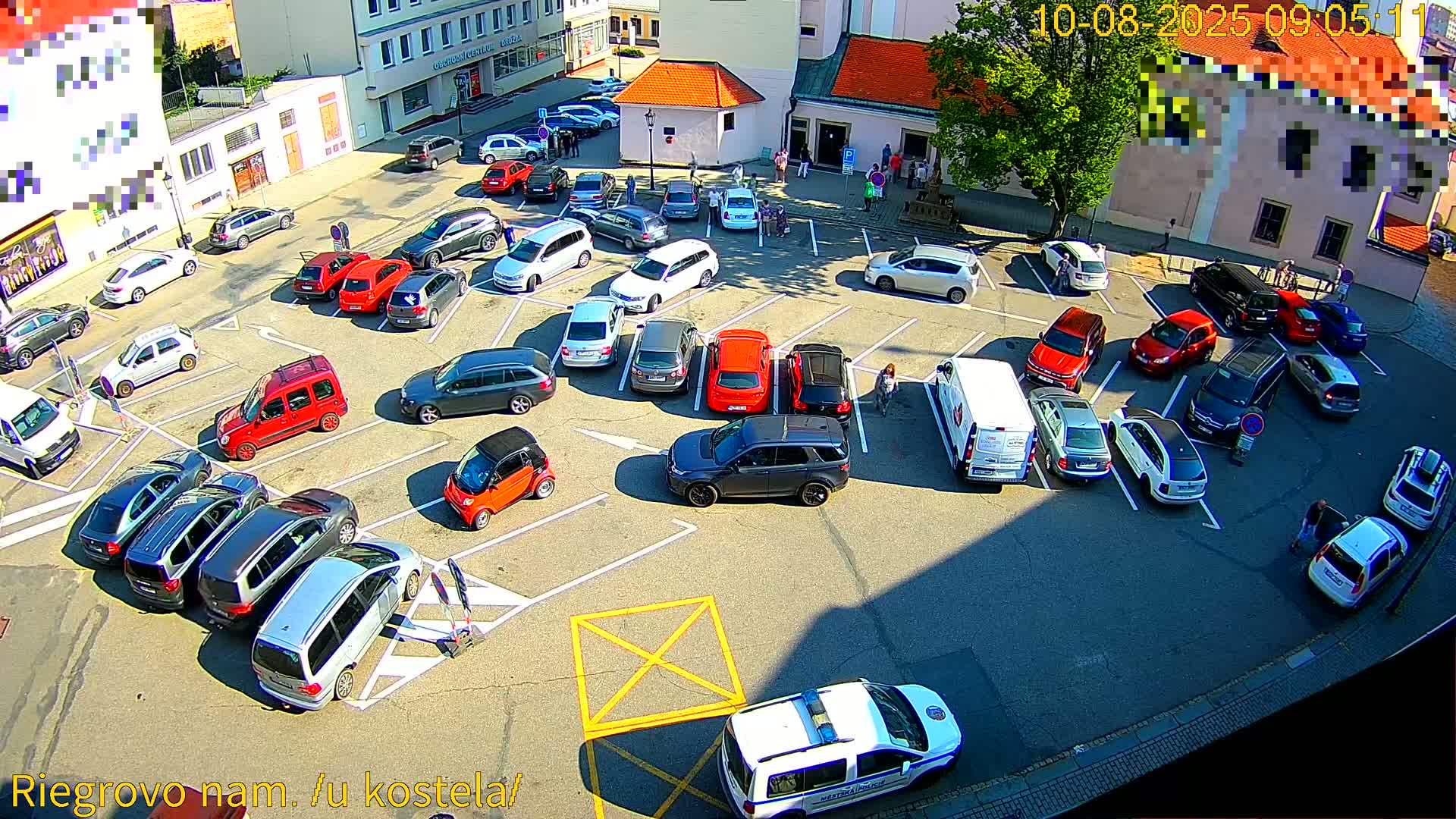 A sunny day shows numerous cars parked in a town square, with a few pedestrians and a police vehicle.