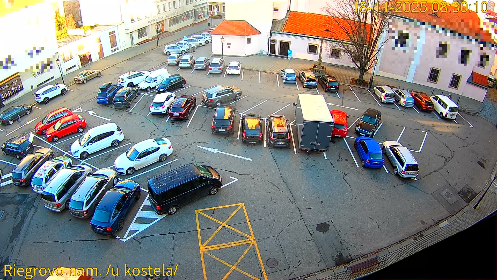 A sunny day shows numerous cars parked in a town square, with a few pedestrians and a police vehicle.
