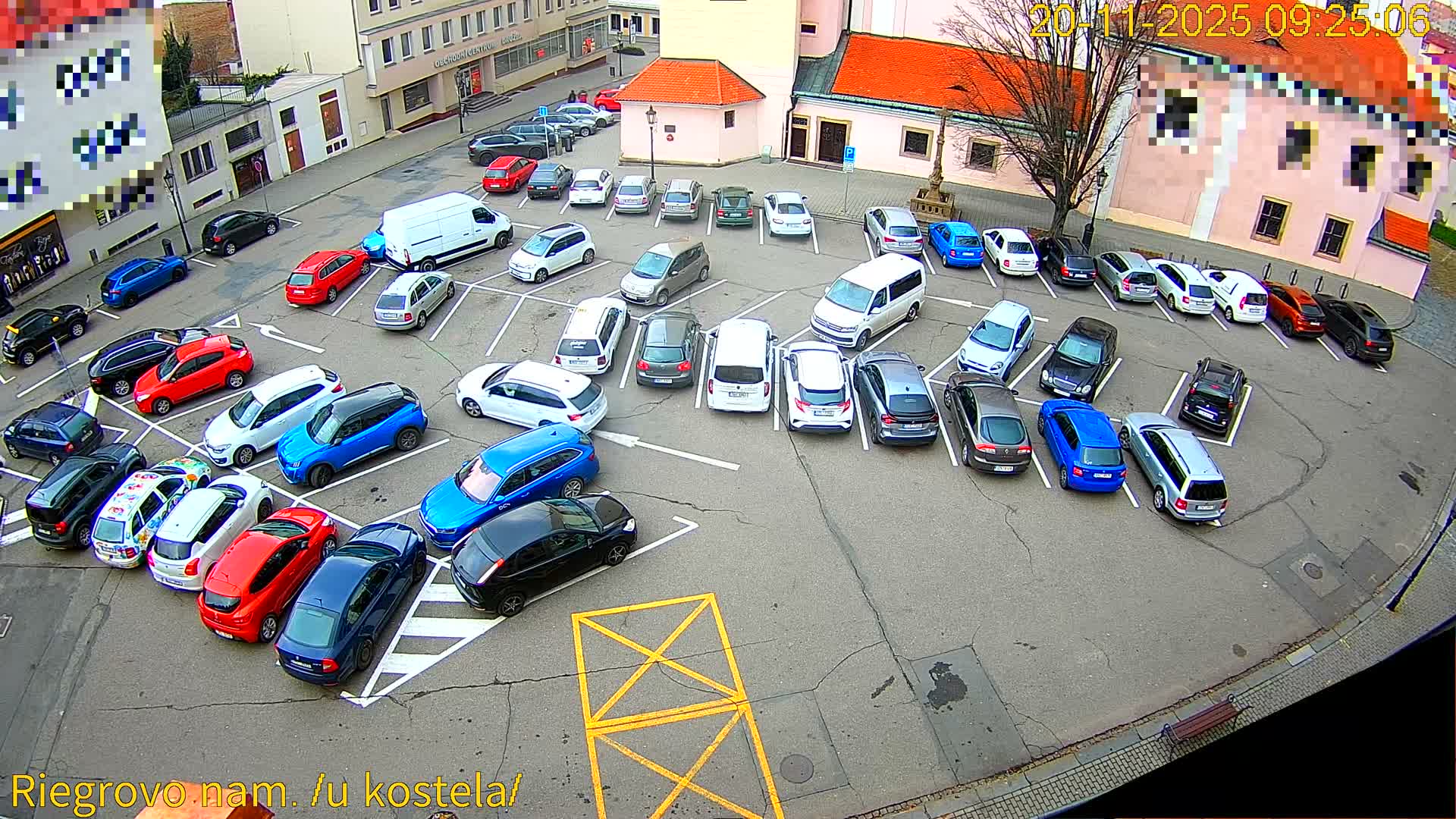 An overcast day illuminates a large outdoor parking lot densely filled with numerous cars of various colors and models, surrounded by buildings with mostly red-tiled roofs and a prominent bare tree.