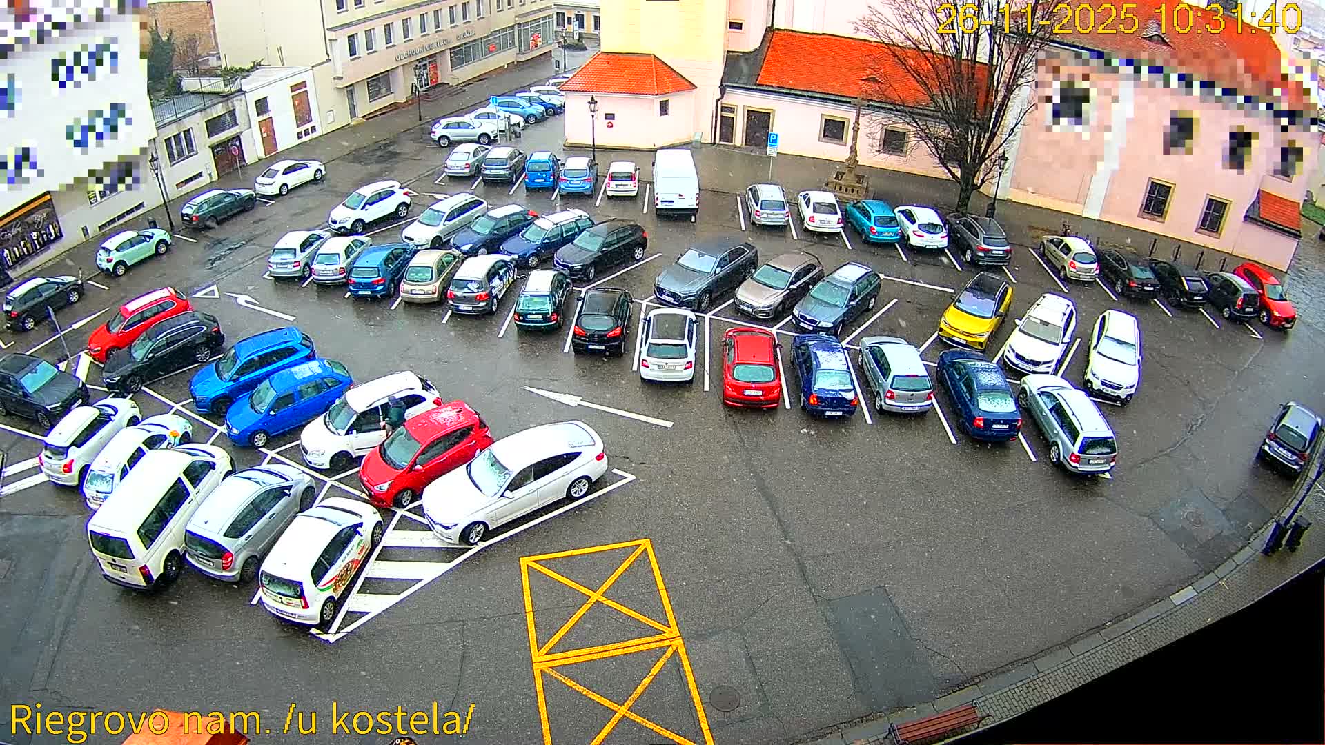 An overhead view captures a large, crowded outdoor parking lot filled with numerous cars of various colors, surrounded by buildings, under an overcast sky with light snow or sleet visibly falling and wetting the ground.