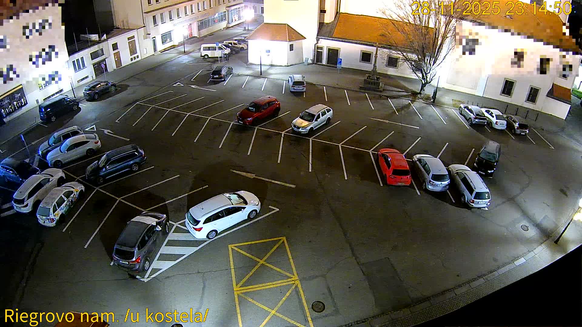 An aerial view captures a brightly lit outdoor parking lot filled with numerous cars at night, bordered by buildings and a bare tree, under clear and dry weather conditions.