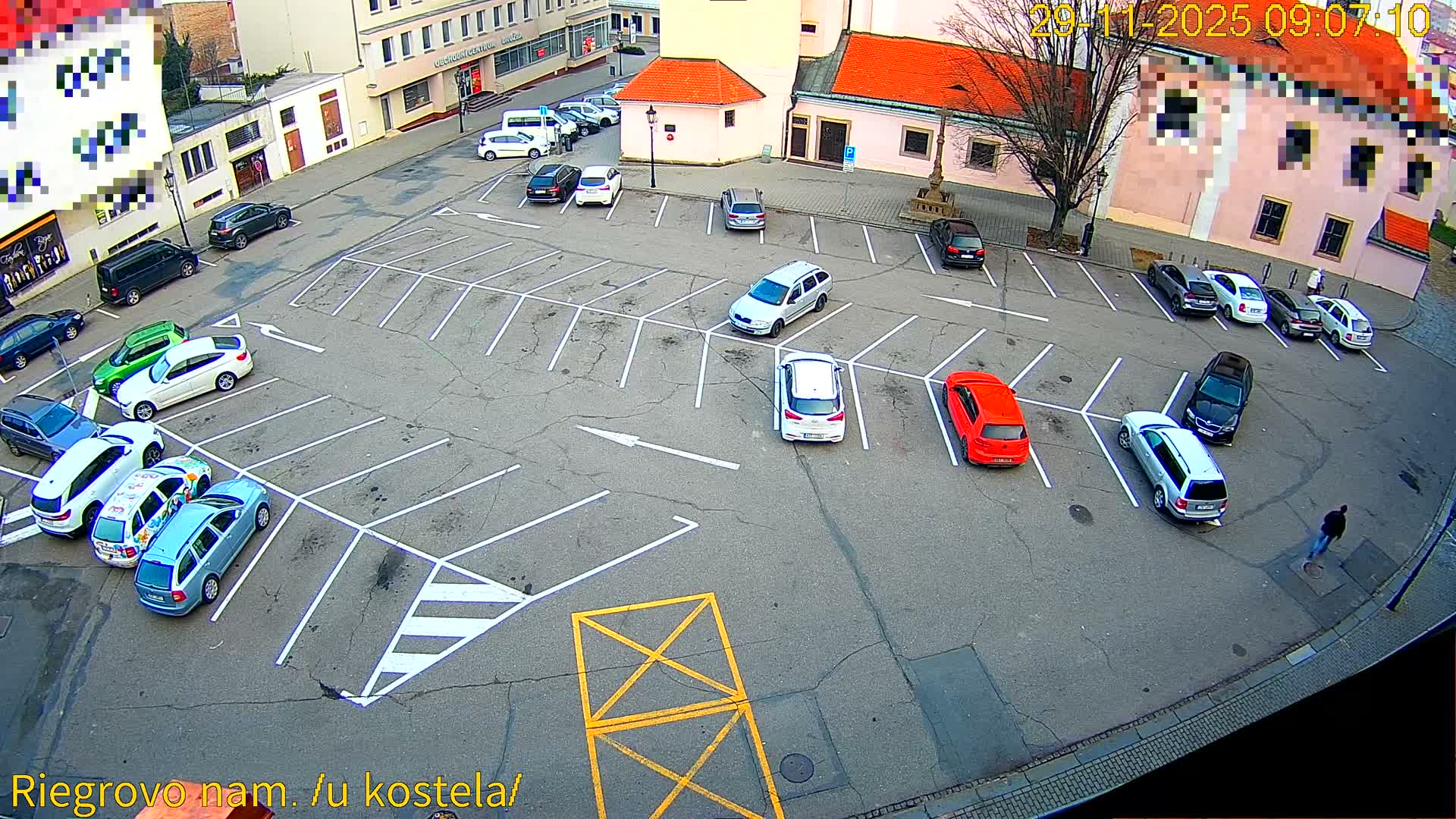 An aerial view reveals an expansive outdoor parking lot filled with various cars and a pedestrian, surrounded by urban buildings with red-tiled roofs, all under a cool, overcast morning sky.