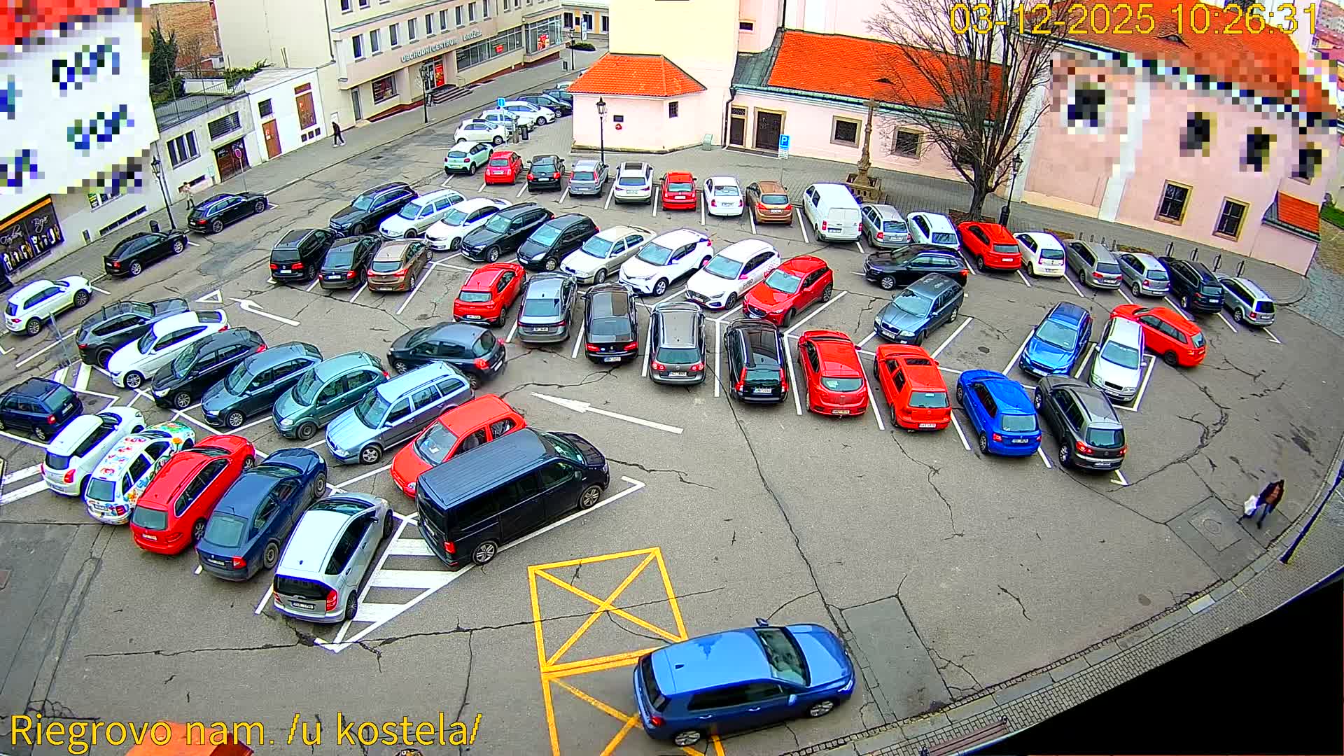 An elevated view captures a large outdoor parking lot densely filled with dozens of cars of various makes and colors, surrounded by buildings and a prominent leafless tree, all beneath an overcast sky.