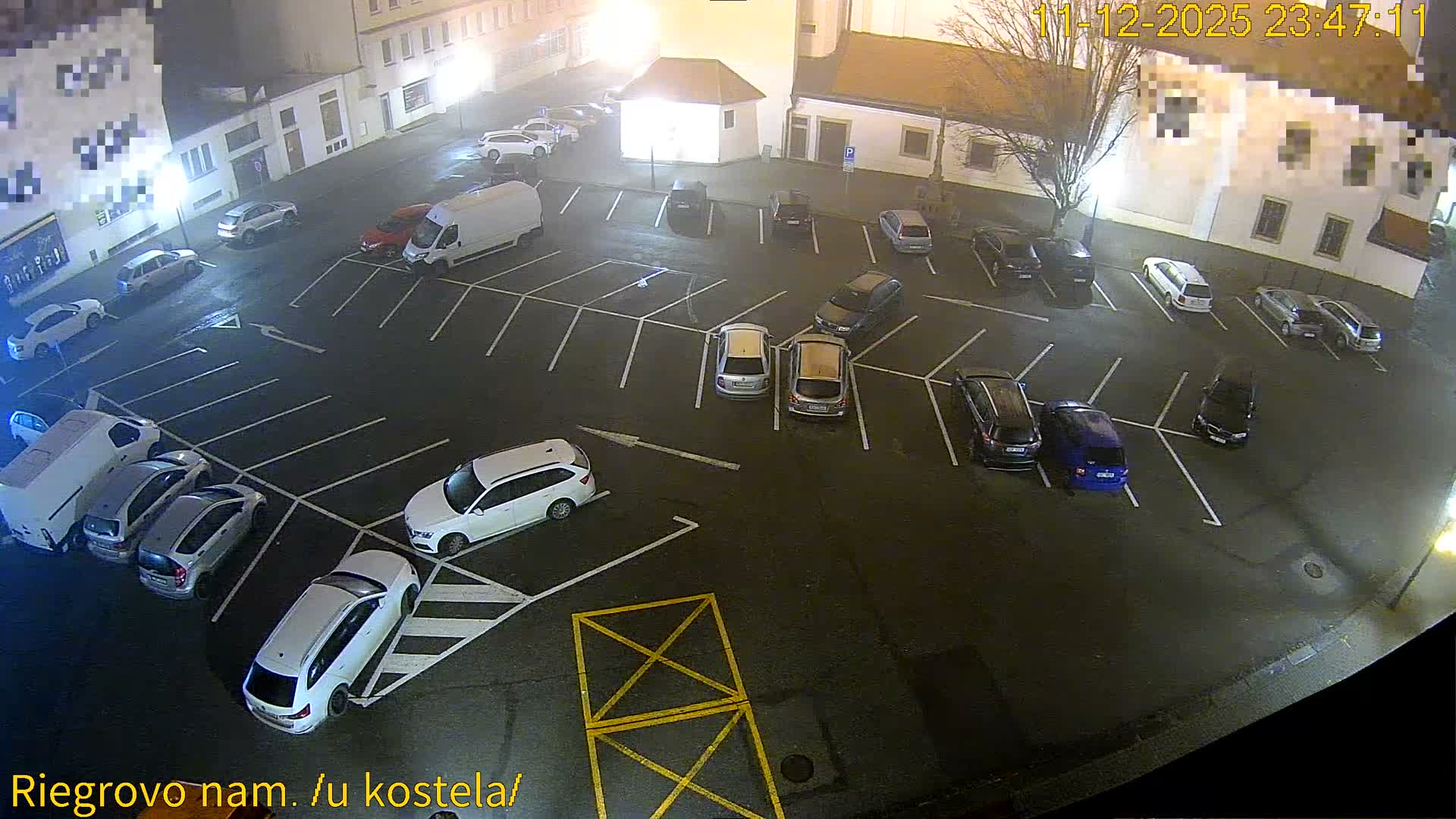 A damp urban parking lot is seen at night, crowded with various cars under a thick fog that blurs the surrounding illuminated buildings and streetlights.