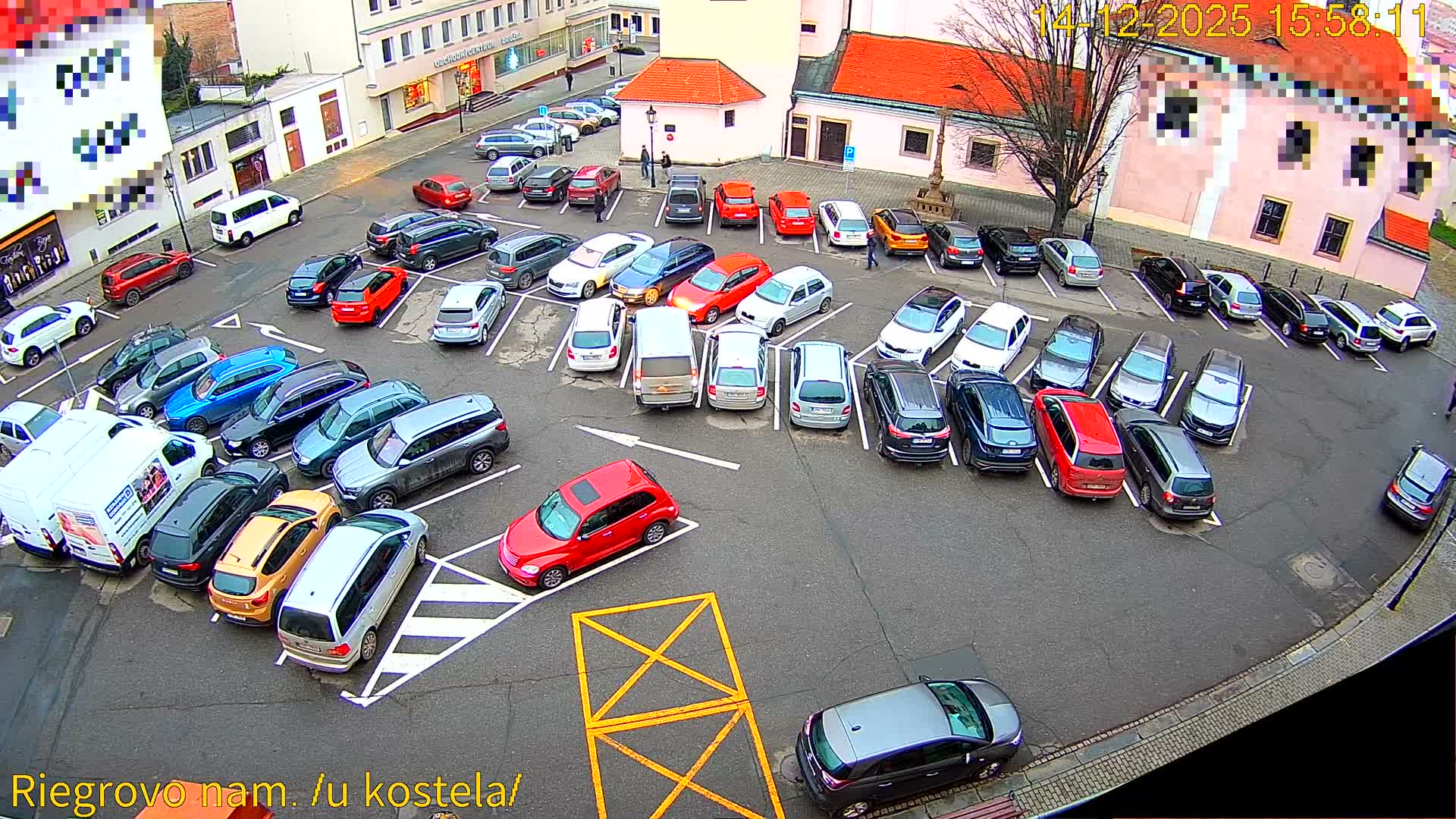 A damp urban parking lot is seen at night, crowded with various cars under a thick fog that blurs the surrounding illuminated buildings and streetlights.