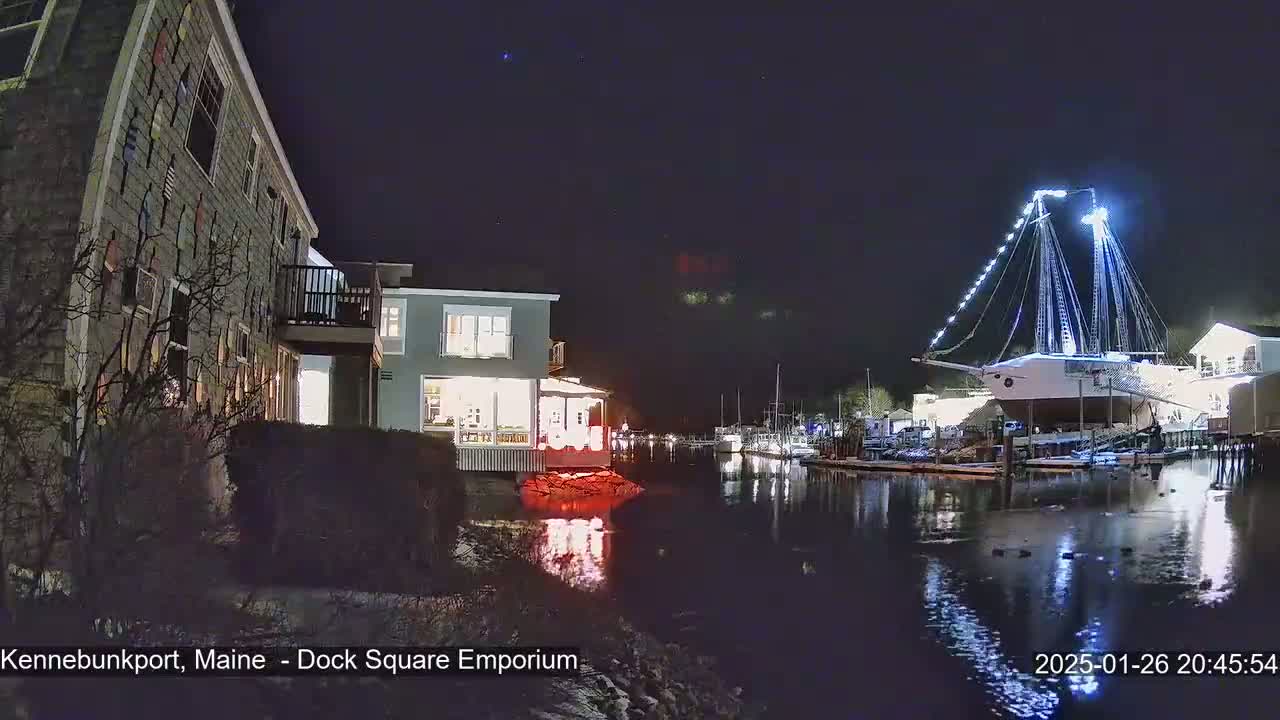 At night, a waterfront scene shows a lit-up building next to a calm body of water where a large, illuminated sailboat is docked among other boats.
