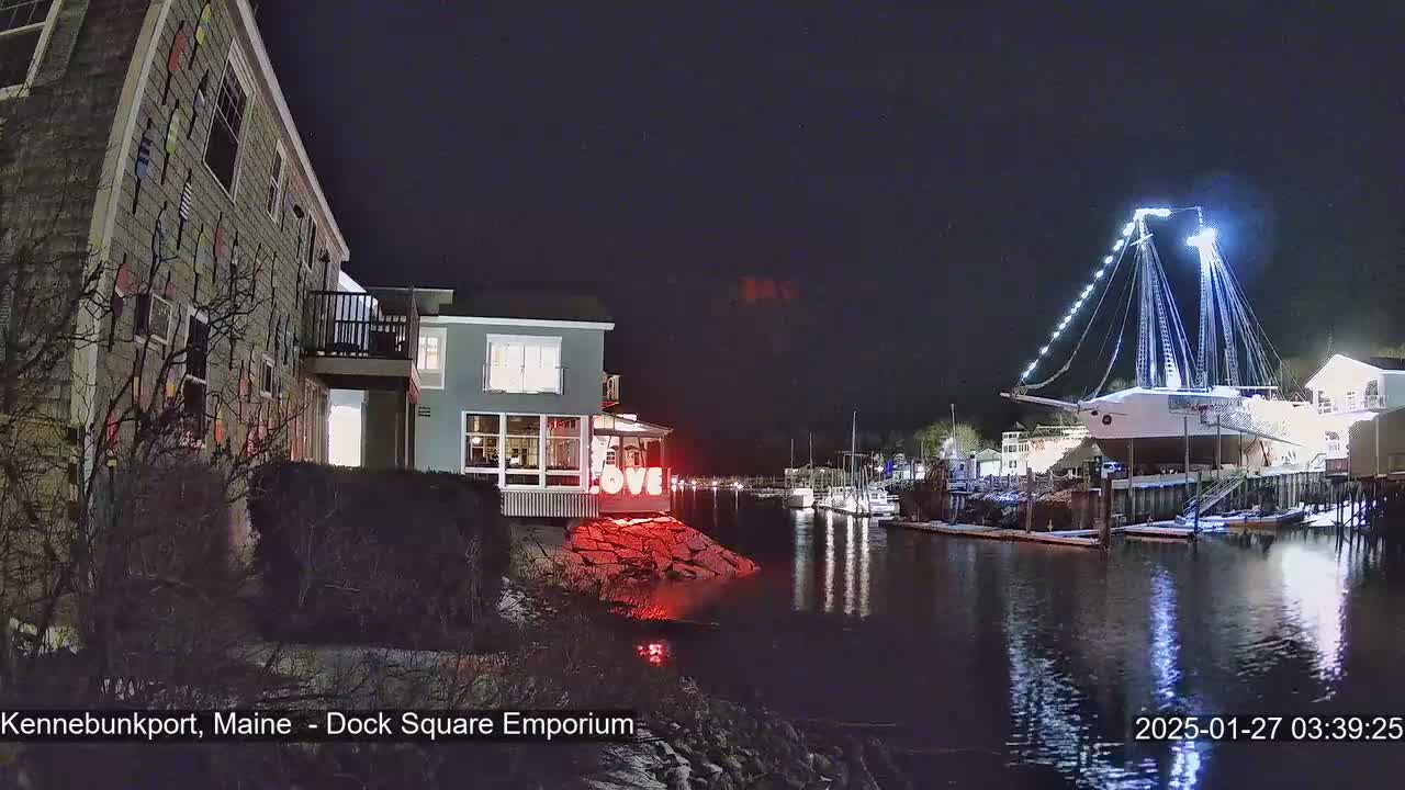 A nighttime view of a harbor with a large, illuminated sailboat docked, a building decorated with flags, and a red "LOVE" sign, under a clear, dark sky with some snow on the ground.