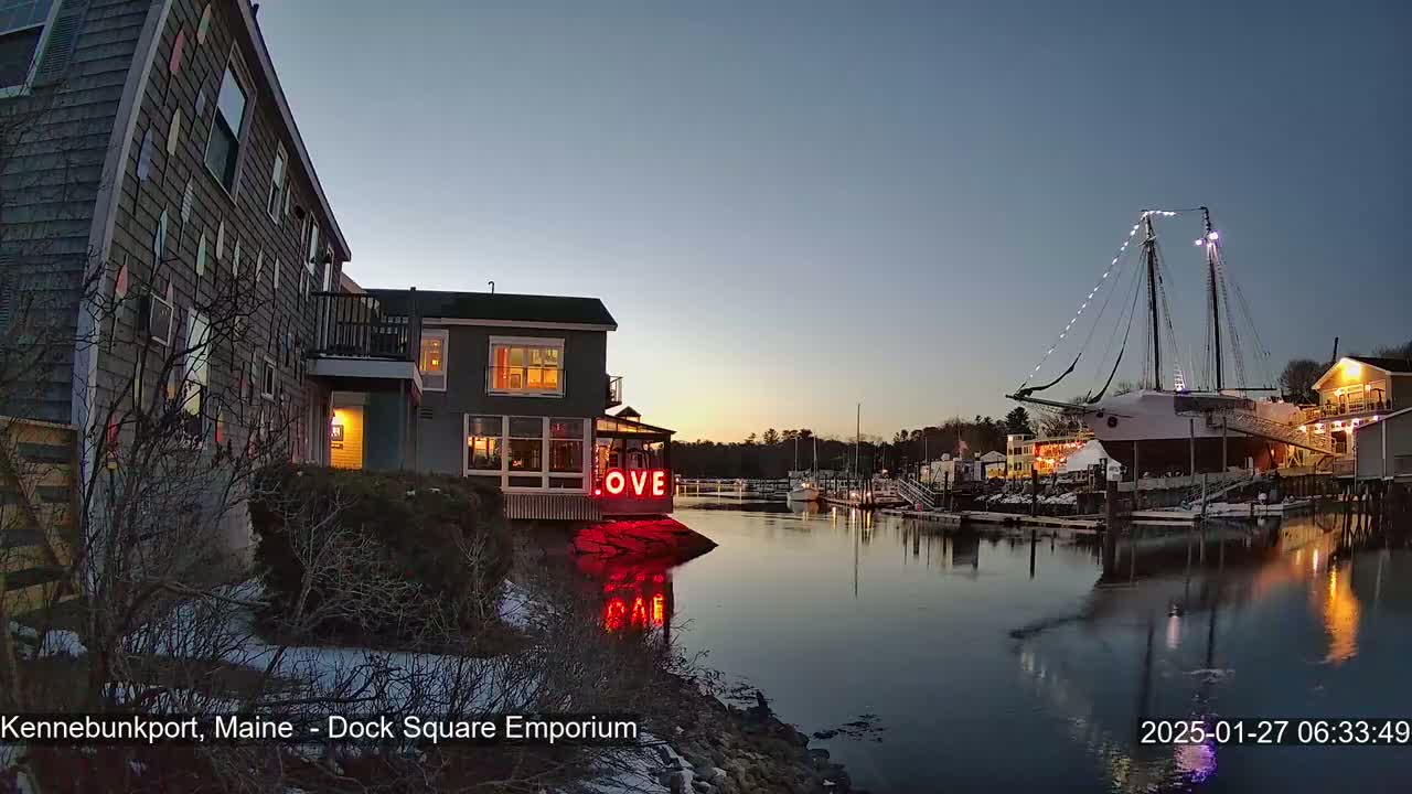 A twilight view of a harbor, featuring a building with illuminated "LOVE" signage, a large sailing ship docked nearby, and snow on the ground.