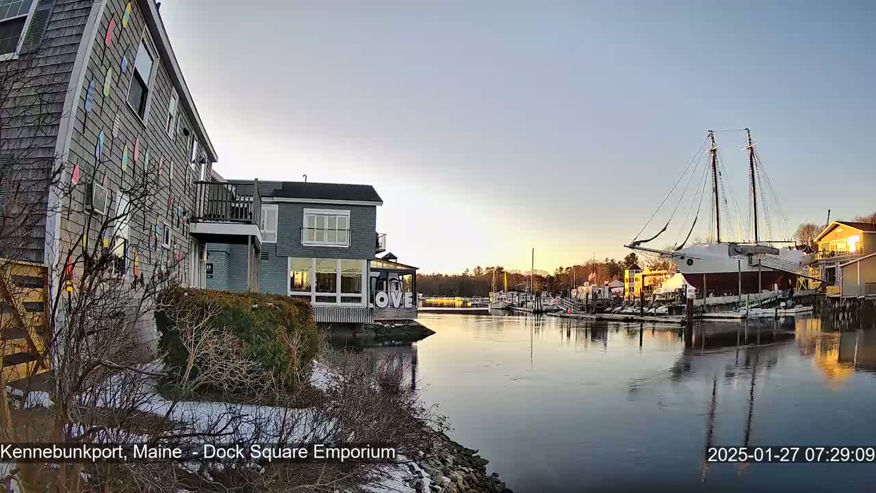 A waterfront scene at sunset shows a building with colorful decorations, other buildings, a large sailboat docked, and a partially frozen waterway with snow on the ground.