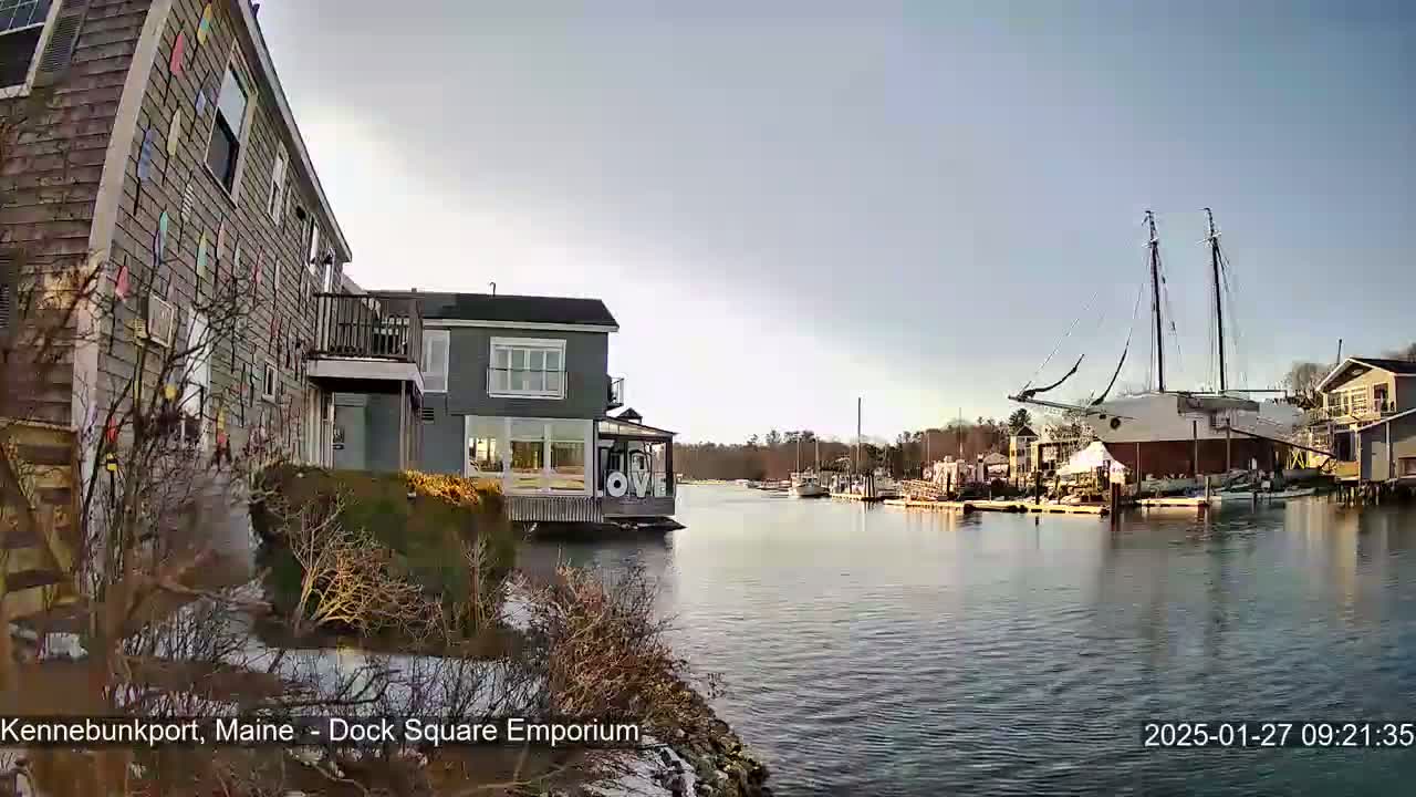 A calm waterway with docked boats, including a large sailing vessel under construction, is seen from the shore near buildings with a partially snowy shoreline.