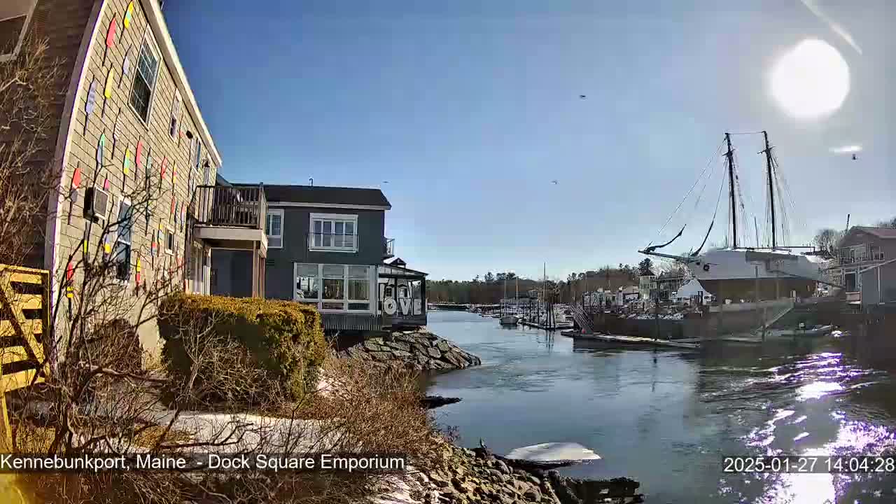A sunny day reveals a waterfront scene with colorful buildings, a docked sailboat, and a calm river reflecting sunlight.