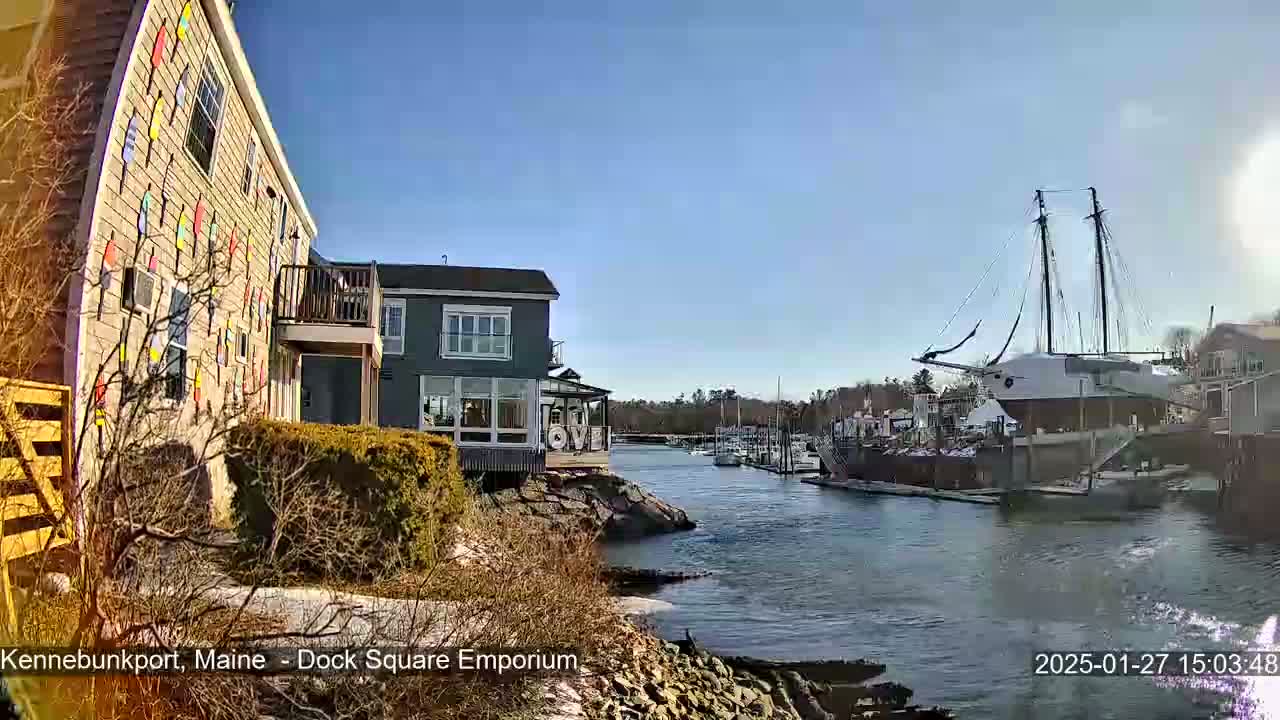 A waterfront scene on a sunny day shows a building decorated with colorful buoys, other buildings, and a large sailboat docked in a calm harbor.