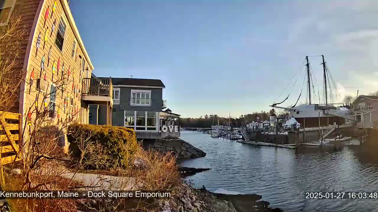 A waterfront scene on a sunny day shows a building decorated with colorful buoys, other buildings, docked boats, and a large sailing ship in a calm harbor.