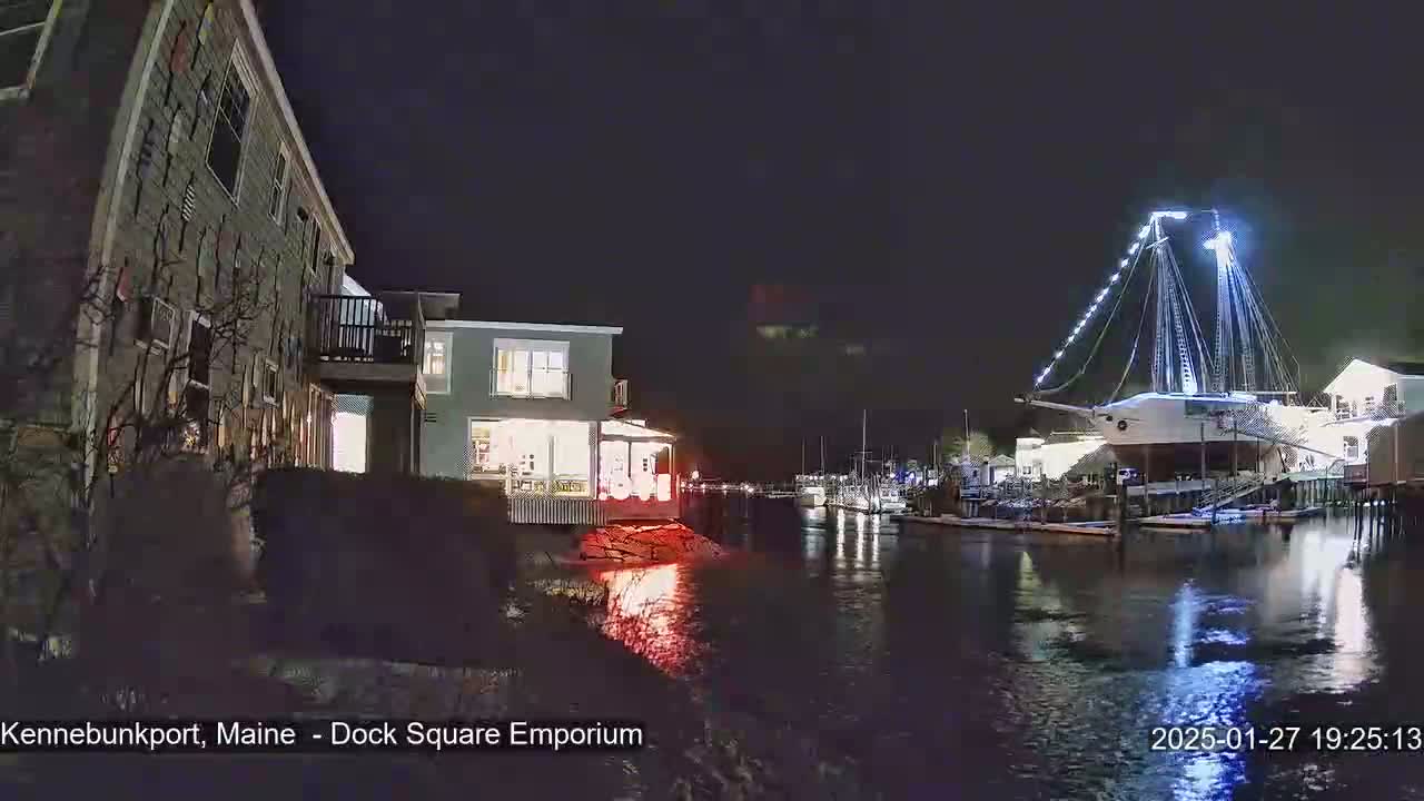 At night, a large illuminated sailboat is docked in a calm harbor next to lit buildings and other boats.