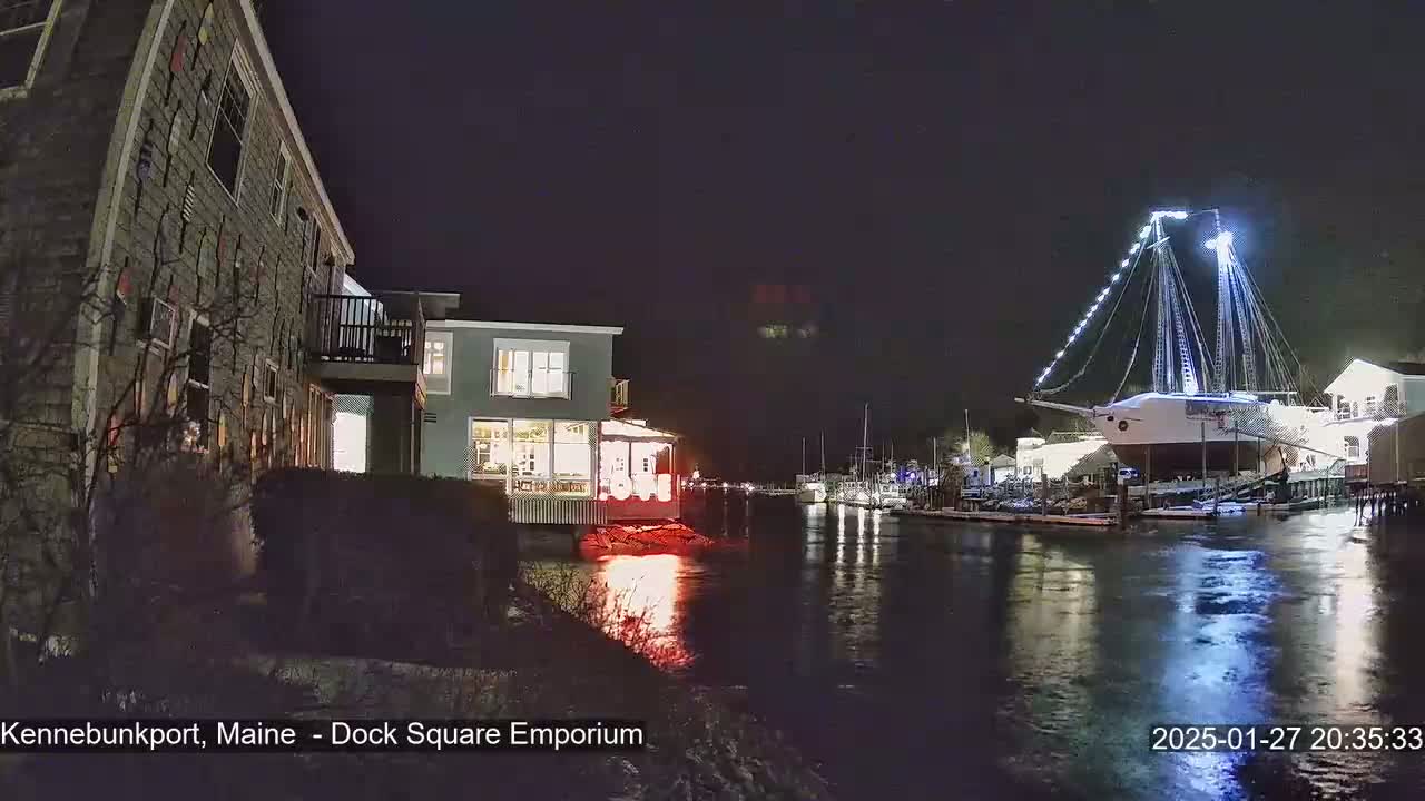 A nighttime view of a waterfront scene shows a docked, illuminated sailing ship and buildings along a calm river.
