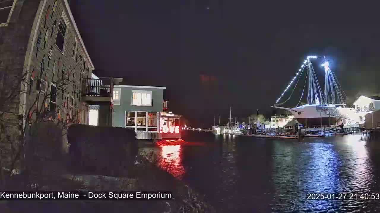 A large illuminated ship is docked at a waterfront at night, next to buildings with lights on, under a clear, dark sky.