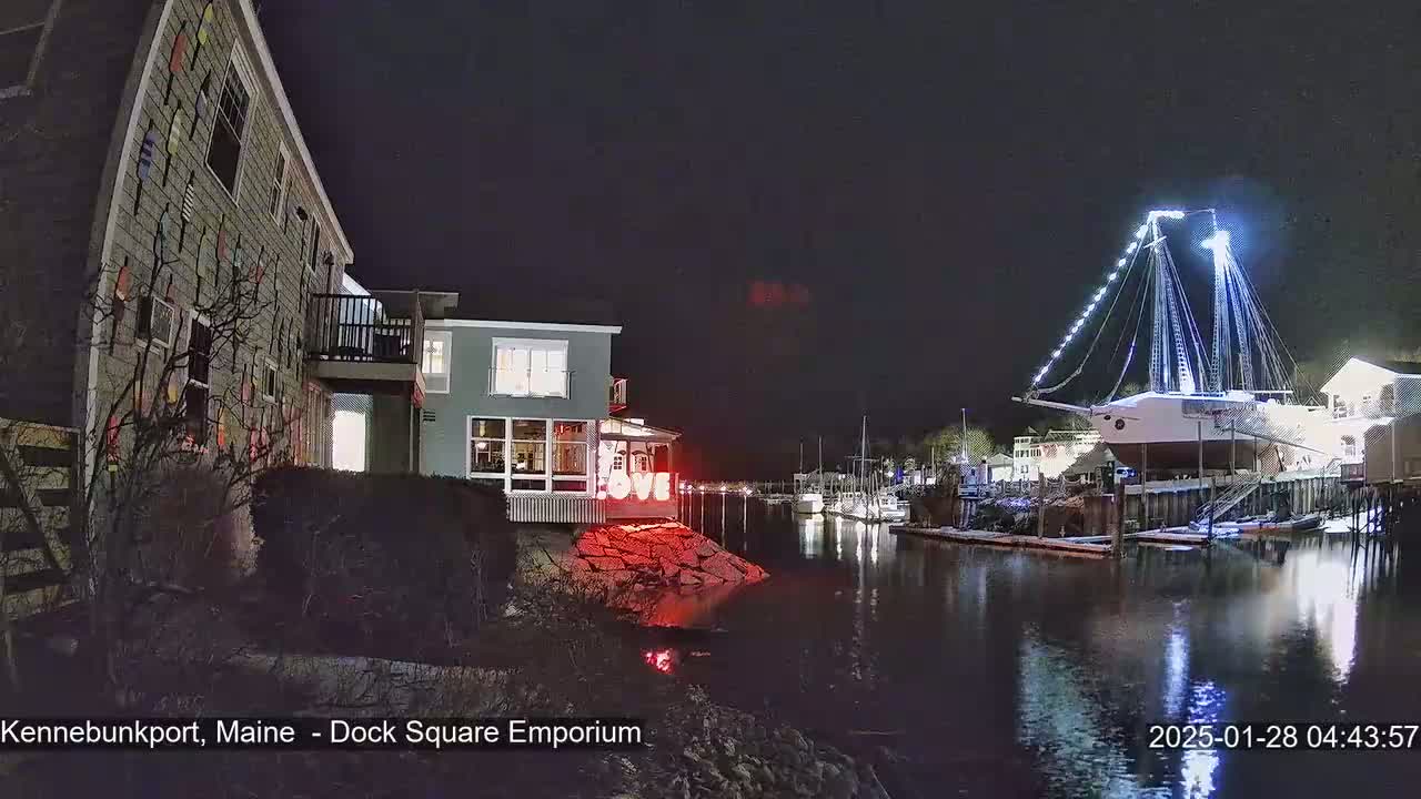 A nighttime waterfront scene shows a building decorated with nautical-themed artwork, a large illuminated ship docked nearby, and calm water reflecting the lights.