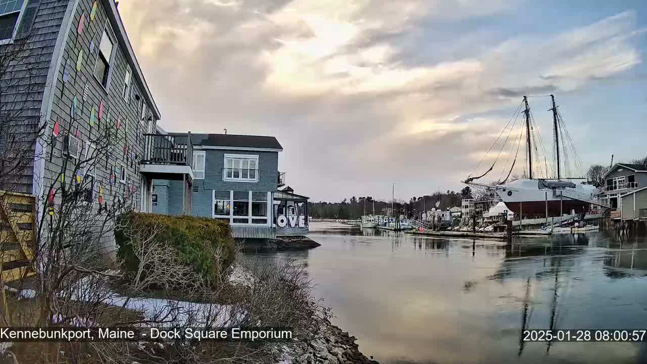 A calm waterway reflects a cloudy sunset sky, with buildings and a large sailboat docked along its banks, some snow visible on the ground.