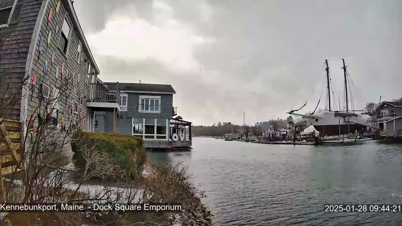 A waterfront scene on an overcast day shows a building with colorful decorations, another building with large windows, a docked sailing ship, and a calm waterway with patches of snow on the ground.
