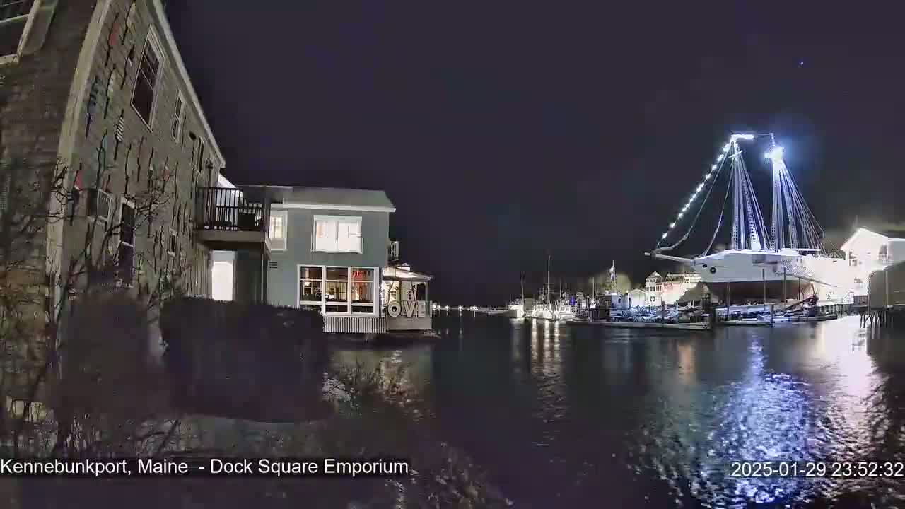 A nighttime view of a harbor with a large, illuminated sailing ship docked, and several smaller boats nearby, with buildings along the shore.