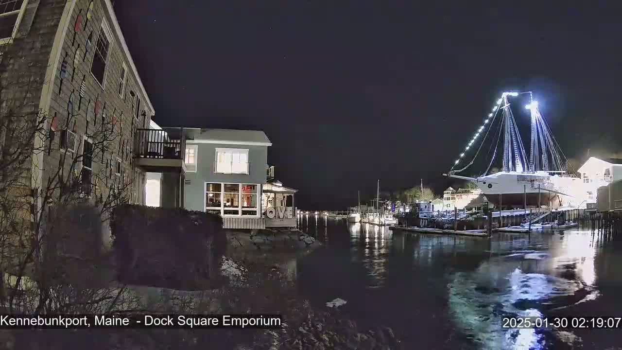 A nighttime view of a harbor with a large, illuminated ship docked, alongside buildings and other boats, under a clear, dark sky with some light snow on the ground.