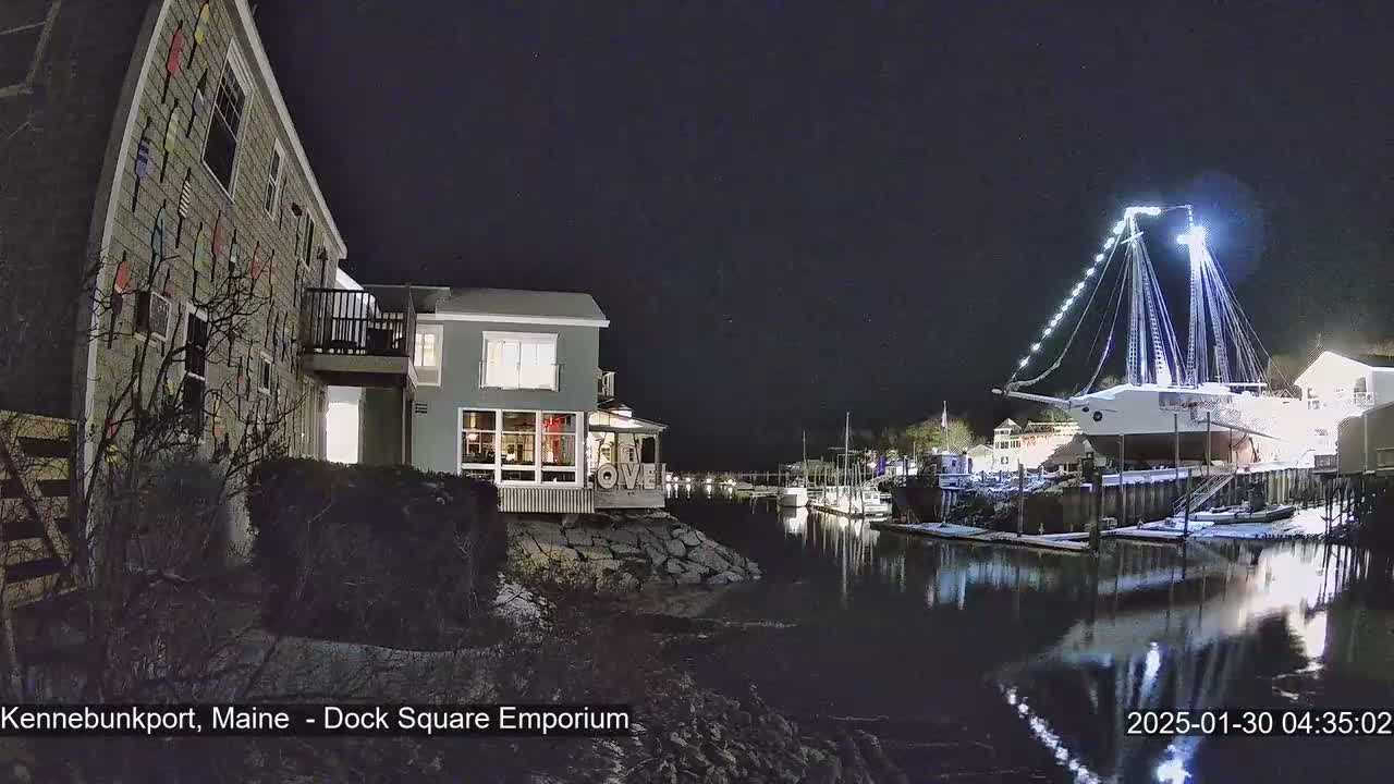 A large, illuminated ship is docked at a waterfront at night, with a building decorated with colorful buoys visible on the left, and snow on the ground near the water.