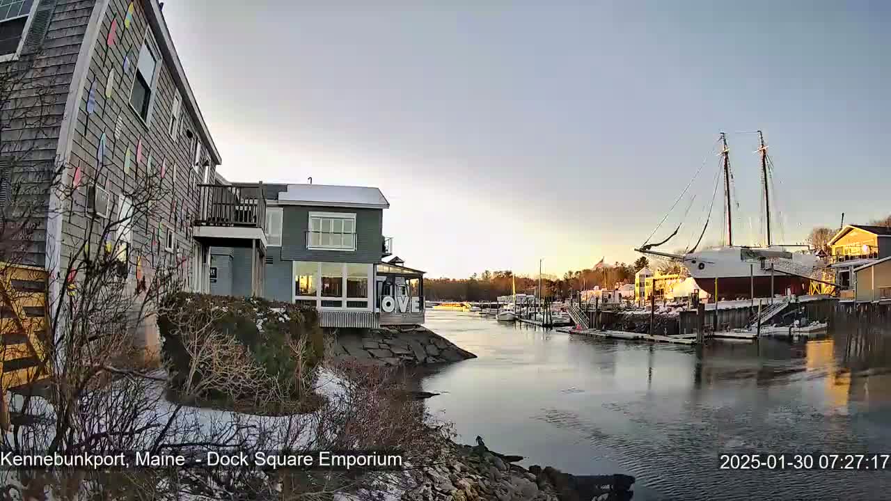 A waterfront scene at sunrise shows a harbor with a large sailing ship docked near buildings, some of which have snow on the ground.