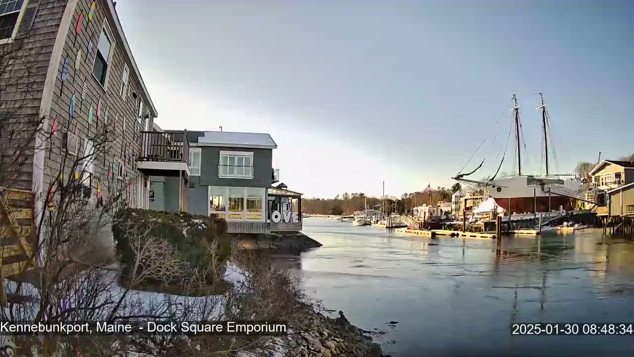 A waterfront view on a partly sunny day shows a large sailboat docked near a building with a "LOVE" sign, other buildings, and some snow on the ground.